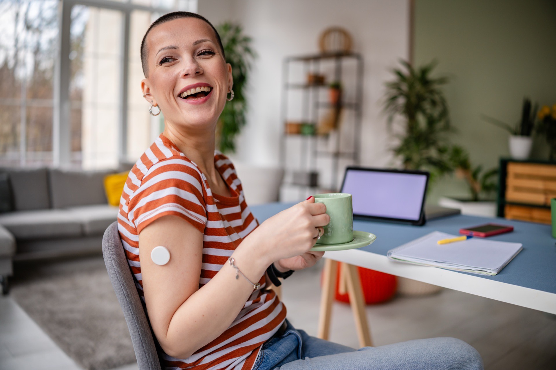 A smiling person with a continuous glucose monitor on their arm, relaxing with a coffee at home.